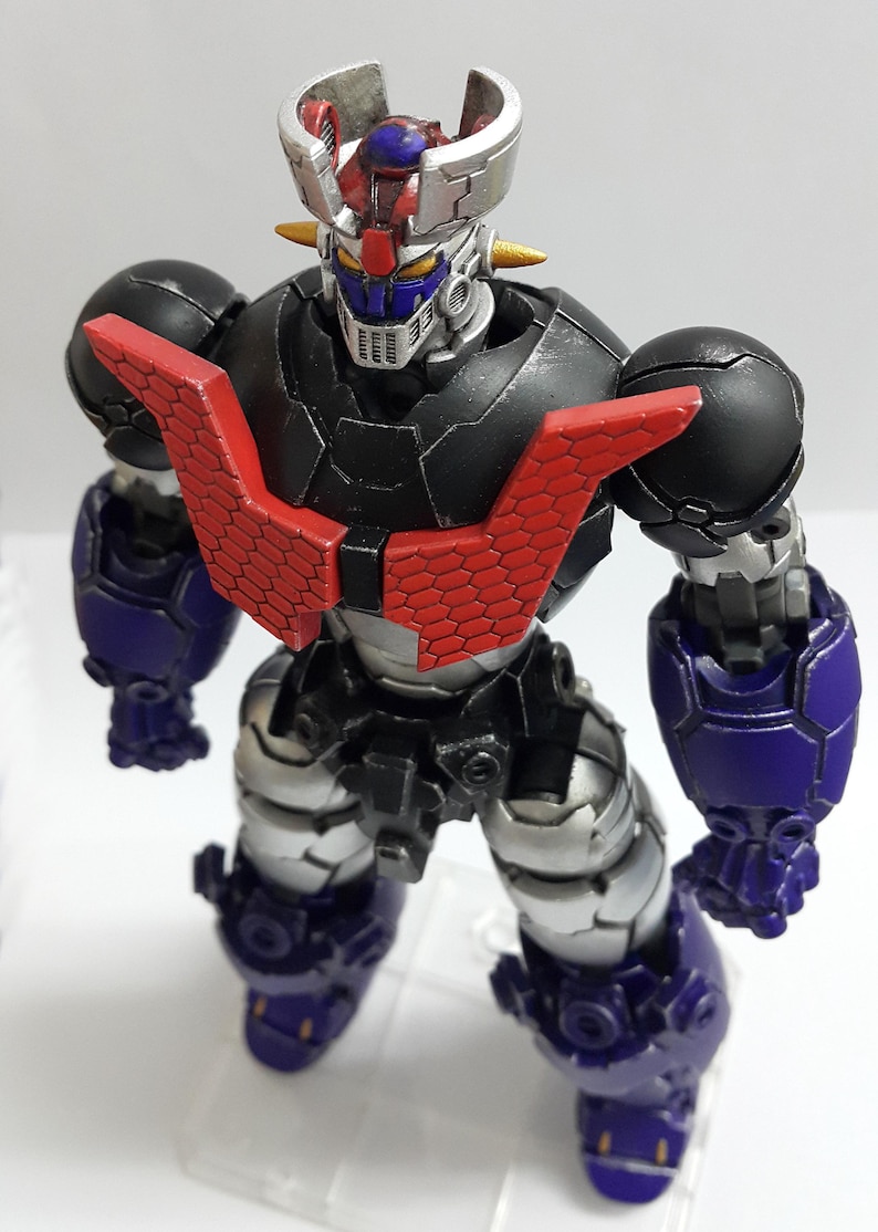 May include: A robotic action figure featuring a black torso with a red hexagonal chest plate, silver limbs, and purple forearm and leg armor. The head has a silver crest, yellow eyes, and a blue face mask, standing on a clear plastic base.