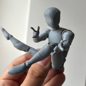 May include: A grey articulated action figure with a smooth, matte finish, featuring a masked head with subtle web-like patterns, a slender build, and multiple joints at the shoulders, elbows, hips, and knees, posed in a dynamic seated position.