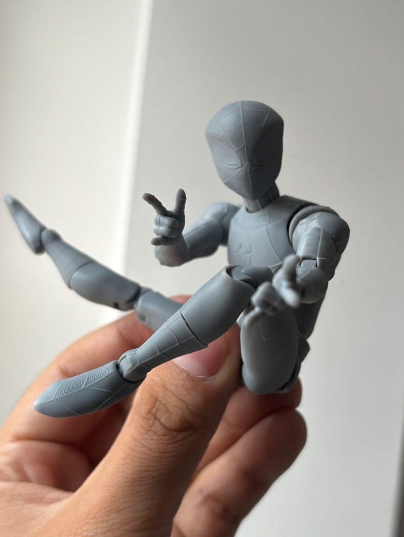 May include: A grey articulated action figure with a smooth, matte finish, featuring a masked head with subtle web-like patterns, a slender build, and multiple joints at the shoulders, elbows, hips, and knees, posed in a dynamic seated position.