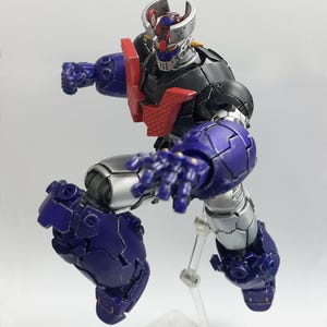 May include: A posable robot action figure featuring a black, silver, and purple color scheme with a prominent red chest plate. The figure has a metallic finish, a horned head design, and is captured in a dynamic mid-air pose with one arm extended.