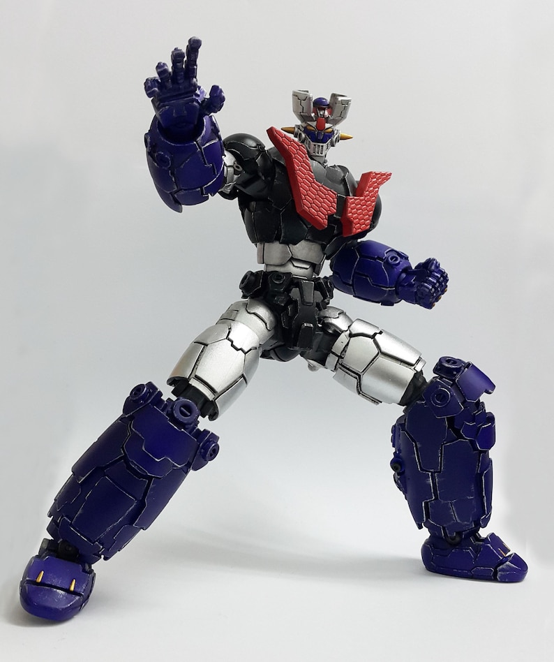 May include: A posable robot action figure featuring a dark blue and silver armored body with a prominent red chest plate. The figure has a detailed head with a silver crest, a black torso, and articulated limbs posed in a dynamic stance.