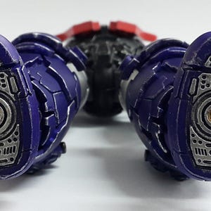 May include: A robotic action figure showing the soles of two purple and silver feet. The feet feature intricate mechanical detailing with circular gold-colored centers and textured metallic plating. The legs are segmented with purple armor.
