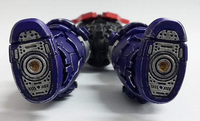 May include: A robotic action figure showing the soles of two purple and silver feet. The feet feature intricate mechanical detailing with circular gold-colored centers and textured metallic plating. The legs are segmented with purple armor.
