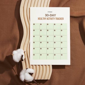 May include: A white 30-day healthy activity tracker with a grid of boxes for tracking fitness goals. The text "Fitness" and "30-Day Healthy Activity Tracker" are printed at the top. The tracker is placed on a wooden decorative piece with cotton stems.