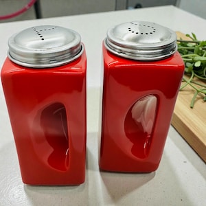 Puede incluir: Un par de saleros y pimenteros rojos de cerámica con tapas de acero inoxidable. Cada salero tiene un cuerpo rectangular con un recorte curvo y agujeros en la tapa.