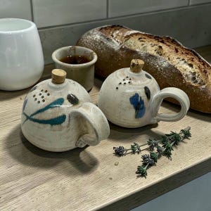 May include: Two small, handmade ceramic teapots with cork stoppers and handles. One teapot has blue and teal painted accents, the other has blue accents. A loaf of bread, a small cup, and a white ceramic cup are in the background.