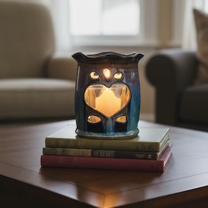 May include: A ceramic wax warmer with a glossy blue and brown glaze, featuring a heart-shaped cutout and a scalloped top edge. A small glass votive holder containing a lit white candle sits inside the center of the heart-shaped opening.