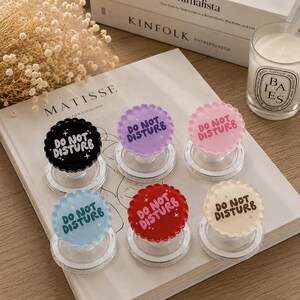 May include: Six circular phone grips with scalloped edges, each featuring the text DO NOT DISTURB and small star icons. The grips come in black, purple, pink, light blue, red, and cream, all mounted on clear, collapsible plastic bases.