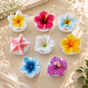 May include: A collection of eight decorative floral and starfish ornaments arranged in a grid. The set includes a yellow and white plumeria, a bright pink hibiscus, a blue and white plumeria, a pink starfish, a white lily, a yellow hibiscus, a blue flower, and a purple bougainvillea, each mounted on a clear circular base.