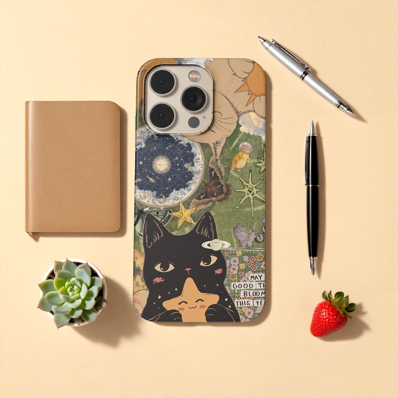 May include: A phone case with a whimsical design featuring a black cat holding a star, surrounded by celestial and botanical illustrations. The case is next to a small succulent, a notebook, pens, and a strawberry.