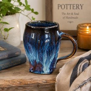 May include: A ceramic mug featuring a glossy glaze with a deep blue and black drip pattern that transitions into a lighter blue at the base. The mug has a curved body, a wide rim, and a sturdy handle with a brown, textured finish on the exterior.