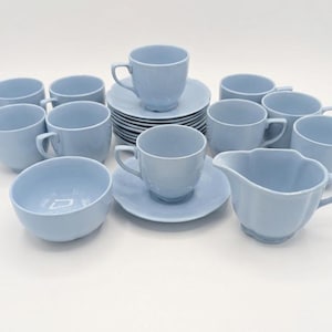 Pode incluir: Um conjunto de chá de cerâmica azul claro, incluindo xícaras, pires, um creme e uma tigela. O conjunto apresenta uma pilha de pires e várias xícaras com alças. O creme tem uma borda ondulada. O conjunto está disposto em uma superfície branca.