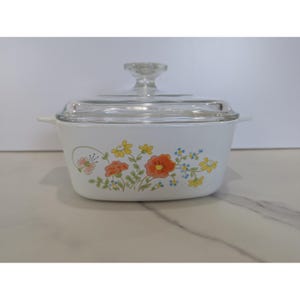 May include: A white rectangular casserole dish with a clear glass lid and integrated side handles. The exterior features a colourful floral pattern with orange, yellow, and blue flowers, green stems, and a small butterfly on the left side.