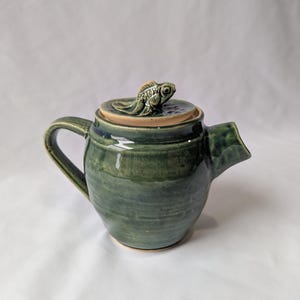 May include: Handmade ceramic teapot in shades of green with a fish-shaped lid handle. The teapot has a curved handle and a spout. The lid is tan with a detailed gold fish. The teapot is a unique piece of pottery.
