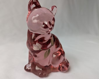 May include: A pink glass cat figurine with painted white flowers and green leaves. The cat is sitting with its ears perked up. The figurine is made of translucent glass.