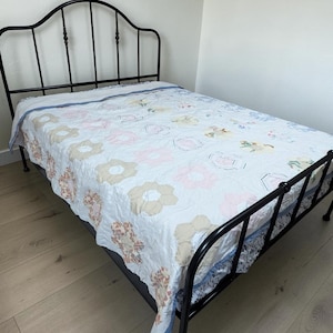 May include: A black metal bed frame with a white quilt featuring a repeating hexagonal floral pattern in soft shades of pink, tan, and blue. The quilt has a light blue border and is draped over the mattress, which rests on the dark metal frame.