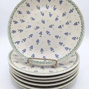May include: A stack of six white ceramic plates with a blue and green floral pattern. Each plate features a repeating design of blue berry clusters and a scalloped green border. The plates are stacked on a gold-colored metal stand.