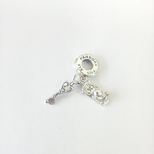 May include: A silver Pandora charm featuring a circular bail stamped with PANDORA, ALE, and S925. Two dangling pendants are attached to the bail: a small ornate key and a rectangular clock-shaped charm with a circular face and detailed markings.