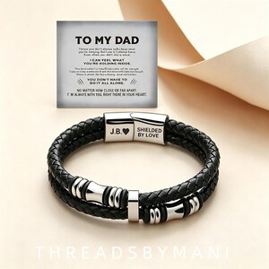May include: A black braided leather bracelet with a silver-toned stainless steel magnetic clasp engraved with J.B. and a heart symbol, and the text SHIELDED BY LOVE. It includes a card with the text TO MY DAD and a sentimental message.