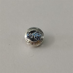 May include: A round silver charm bead featuring the text "we can do anything" engraved along the bottom edge. The center displays a stylized script design encrusted with small, sparkling light blue and dark blue stones set in metal.