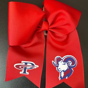 May include: A large red hair bow made of grosgrain ribbon with two long tails. The left tail features a stylized letter P logo in red, white, and blue. The right tail displays a graphic of a ram head in white, red, and blue colors.