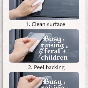 May include: A three-step instructional guide for applying a white vinyl decal to a car window. The decal features the text 'Busy raising feral children' in a gothic font, accompanied by a checkered lightning bolt and small star icons.
