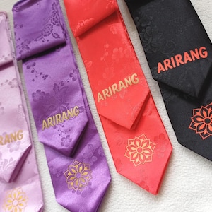 May include: Four neckties in light purple, purple, red, and black, each featuring a tonal floral pattern, the word ARIRANG printed in a contrasting colour, and a stylised flower graphic near the pointed tip of the fabric.