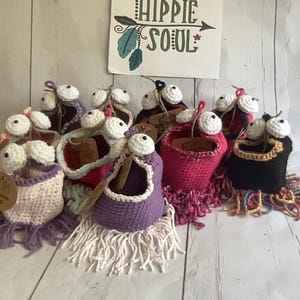 Puede incluir: Una colección de muñecas de ganchillo hechas a mano con grandes ojos blancos y flecos de hilo de colores. Las muñecas son de varios colores, incluyendo morado, rosa y negro. Un letrero en el fondo dice "Hippie Soul".