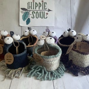 Puede incluir: Una colección de cestas de ganchillo hechas a mano con ojos saltones caprichosos. Cada cesta es de un color diferente, incluyendo azul, marrón y beige, y tiene un fleco desgreñado. Un letrero en el fondo dice "HIPPIE SOUL".