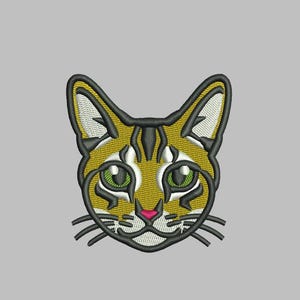 May include: An embroidered design of a cat's face, featuring a detailed pattern in shades of gold, black, and white. The cat's eyes are green, and the nose is pink. The design is 2.52 inches high and 2.34 inches wide. The text "*CAT*" is above the design.