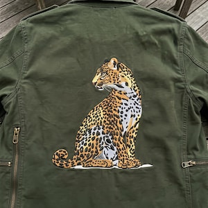 Puede incluir: Chaqueta verde oliva con un diseño de leopardo bordado en la espalda. La chaqueta de estilo militar tiene cuello, bolsillos con cremallera y cierre frontal con cremallera. El leopardo está representado en tonos marrones, negros y crema.