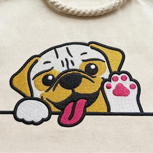 May include: Cream-colored tote bag with a detailed embroidered pug design. The pug has a yellow and white head, black eyes, and a pink tongue and paw. The bag has a rope handle.