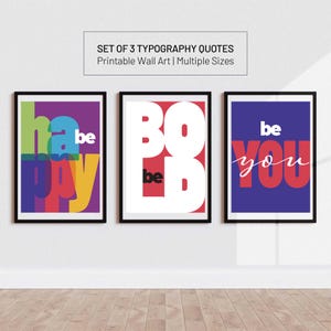 Puede incluir: Conjunto de tres impresiones artísticas tipográficas enmarcadas. Las impresiones presentan las palabras "happy", "bold" y "be you" en fuentes coloridas y superpuestas. Las impresiones se exhiben en una pared blanca con suelo de madera.