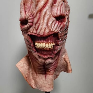 May include: A full-head mask featuring a textured, fleshy, pinkish-red surface with deep folds and creases. The mask displays a wide, open mouth revealing a set of realistic human-like teeth, with thin metal wires wrapped around the cheek areas.