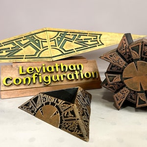 Op de afbeelding: Een goudkleurige en zwarte geometrische puzzeldoos, een driehoekig piramidevormig object en een lang, puntig goudkleurig en zwart decoratief stuk met ingewikkelde geëtste patronen. Een rechthoekige standaard toont de tekst Leviathan Configuration in gele letters.
