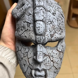 May include: A grey mask with a cracked, stone-like texture. The mask has a prominent nose, fanged mouth, and eye openings. The top of the mask has a textured design. The mask is held by a person.