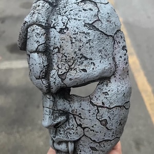 May include: A gray mask with a cracked, stone-like texture. The mask has a detailed face with a prominent nose, fangs, and eye openings. The mask is held in a hand, showcasing its three-dimensional form.