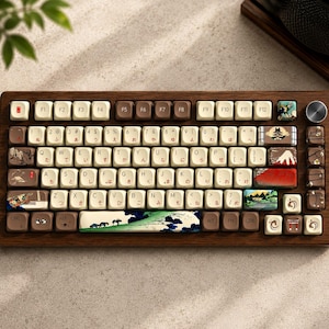 Könnte beinhalten: Eine mechanische Tastatur mit einem dunklen Holzrahmen und einem metallischen Drehknopf. Die Tastenkappen haben ein cremefarbenes und braunes Design mit japanisch inspirierten Landschaftsmotiven, darunter ein Berg und ein Boot.