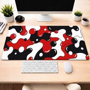 May include: A rectangular desk mat with a bold, abstract design. The pattern features flowing shapes in red, black, and white, creating a modern and eye-catching aesthetic. The mat is placed on a light-colored wooden desk, with a keyboard and mouse visible.