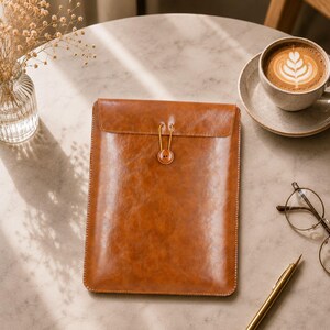 May include: A brown leather tablet sleeve with a button closure. The sleeve is rectangular and has a flap that secures with a button and a loop. The leather has a slightly distressed appearance. A cup of coffee and glasses are on the table.