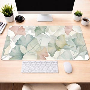 May include: A rectangular desk mat with a watercolour design of ginkgo leaves in pink, green, and cream. The leaves overlap on a white background. A white keyboard and mouse are visible on the desk mat.