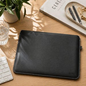 Puede incluir: Una funda para portátil de cuero negro con costuras blancas en los bordes. La funda tiene un diseño minimalista y la marca "quoko" es visible en la esquina inferior derecha. La funda está sobre una superficie de madera.