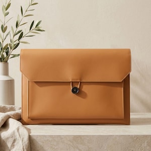 May include: A tan leather document case with a flap closure and a black button. The case is rectangular and has a simple, minimalist design. A small vase with green leaves is in the background.