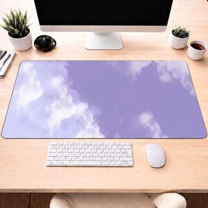May include: A rectangular desk mat with a watercolour design of a cloudy sky in shades of purple and white. A white keyboard and mouse are at the bottom. The mat is on a light wood desk, with a computer monitor and accessories in the background.