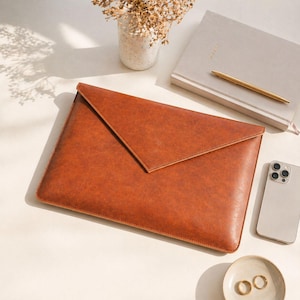 May include: A brown leather laptop sleeve with a flap closure. The sleeve is on a white surface, next to a notebook, pen, smartphone, and small dish with rings. A small vase with dried flowers is also present.