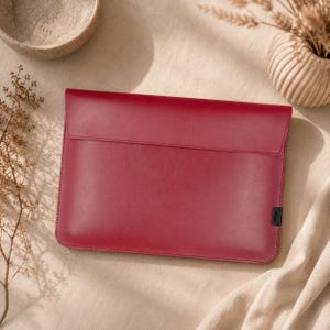 Könnte beinhalten: Eine weinrote Laptop-Hülle aus Leder mit Klappenverschluss. Die Hülle hat ein minimalistisches Design mit einer genähten Bordüre und einem kleinen schwarzen Etikett in der unteren rechten Ecke. Der Hintergrund umfasst einen beigen Stoff und dekorative Elemente.