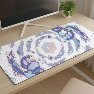 May include: A rectangular white desk mat with a whimsical design. It features a central iridescent geometric shape and paw print, surrounded by blue, purple, and yellow celestial elements and paw prints. The mat is on a wooden desk.