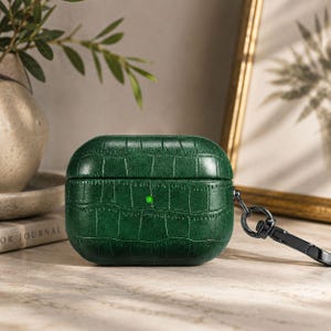 May include: A dark green, textured case for wireless earbuds. The case has a crocodile skin pattern and a black metal carabiner clip. The case is sitting on a light-coloured surface.