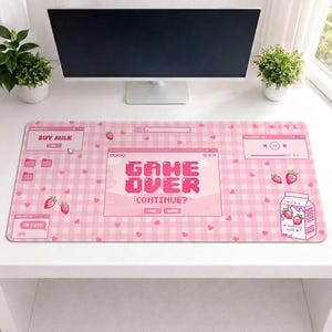 Op de afbeelding: Een roze bureaumat met een ruitpatroon en pixelachtige graphics. De mat bevat de woorden "GAME OVER CONTINUE?" en "BUY MILK". Het bevat ook aardbei- en hartillustraties en een aardbeienmelkpak.