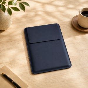 May include: A navy blue tablet sleeve with a flap closure. The sleeve is made of a smooth material and is rectangular in shape. The image is taken from a top-down perspective, with the sleeve resting on a light-colored wooden surface.
